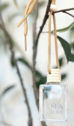 A serene and minimalist aesthetic featuring a square glass bottle labeled "sea love," suspended by a golden rope against a soft-focus backdrop of gentle greenery.