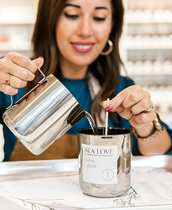 A woman carefully pours wax into a metal container labeled 'sea love' while making a handmade candle.